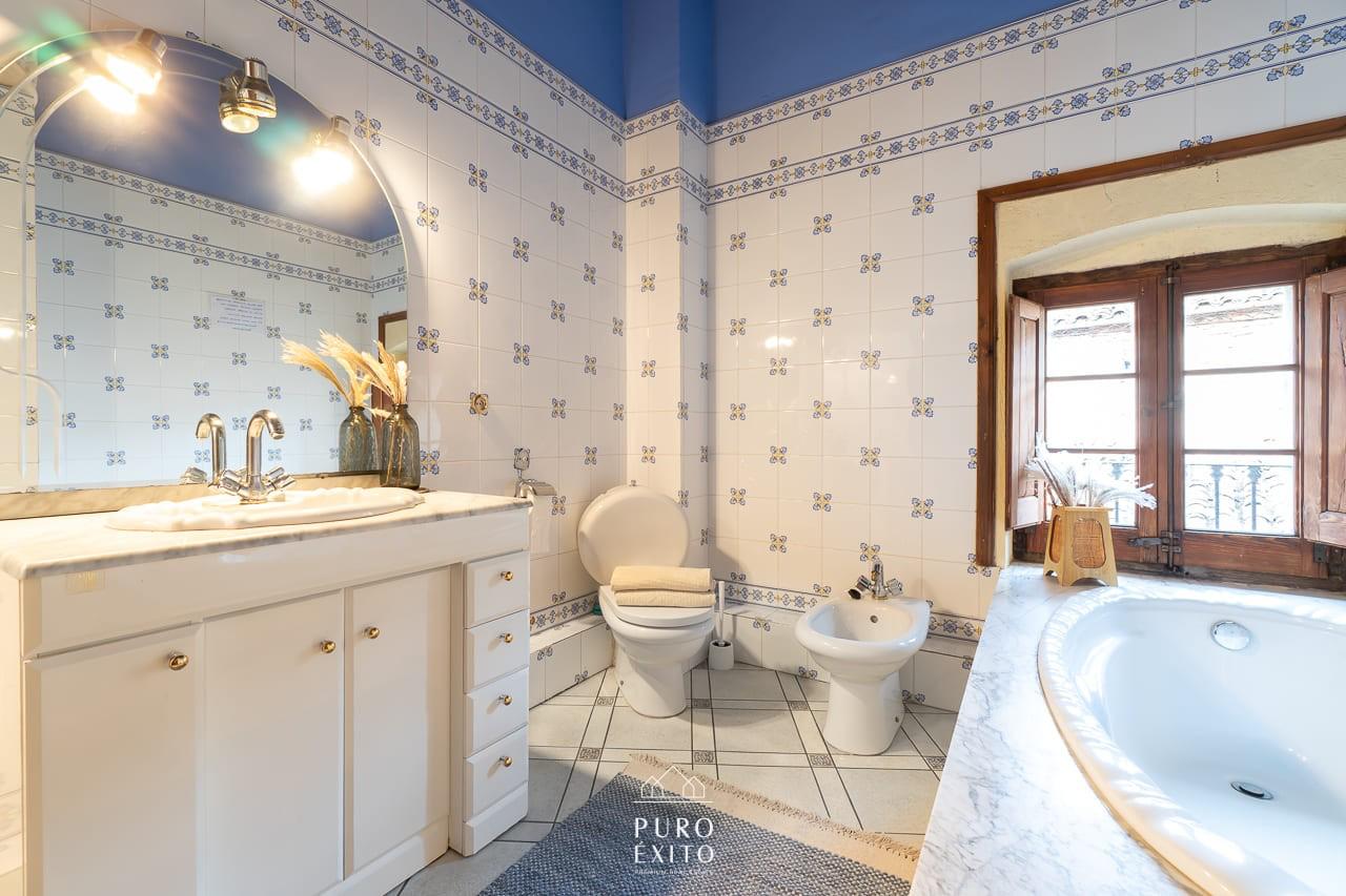 A bathroom tiled in blue and white, with a marble-topped basin unit, a bath set into a marble surround and a shuttered window.