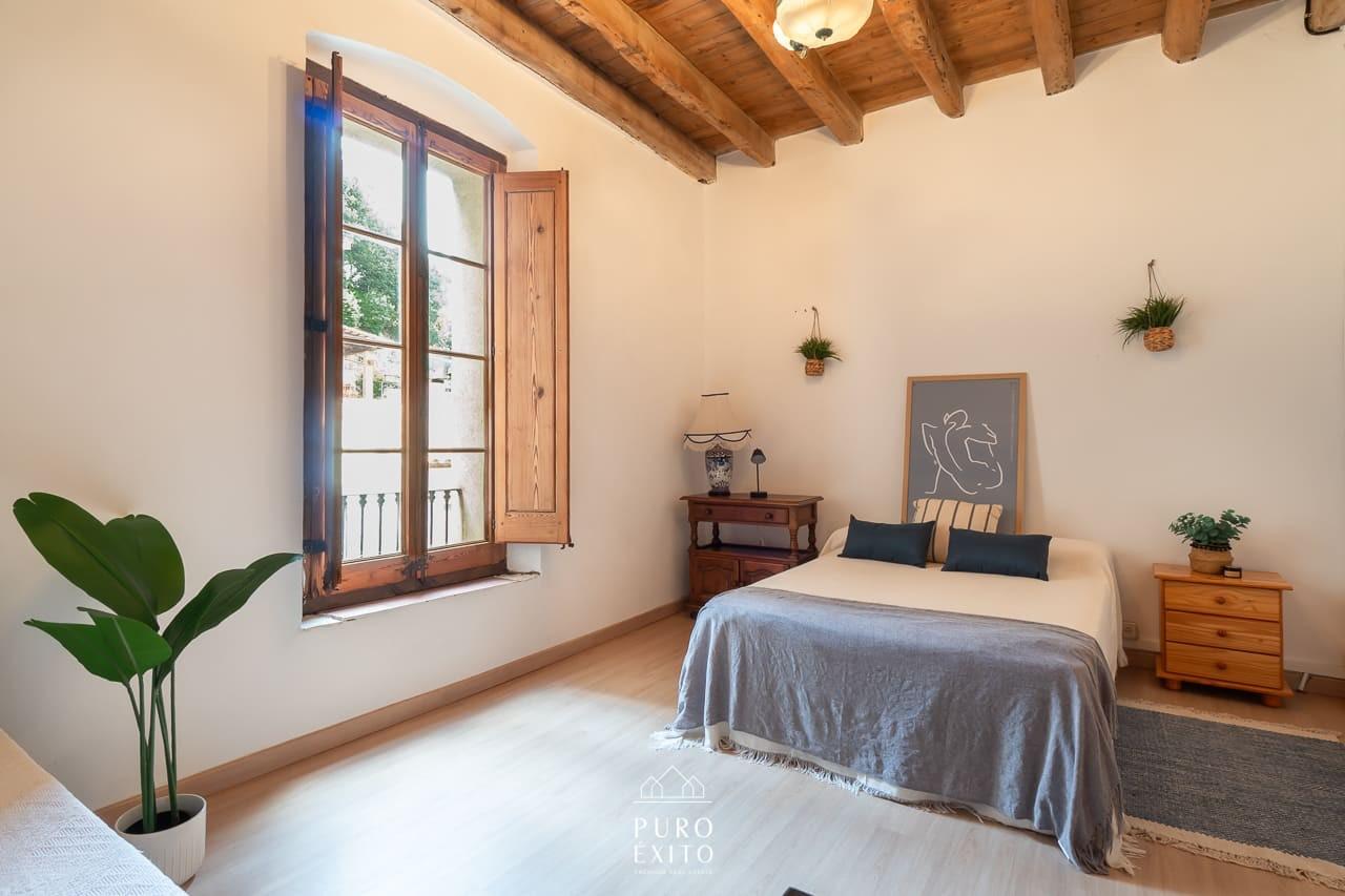 A bedroom with a double bed, pale beamed ceiling and a tall window with wooden shutters open onto a balcony.