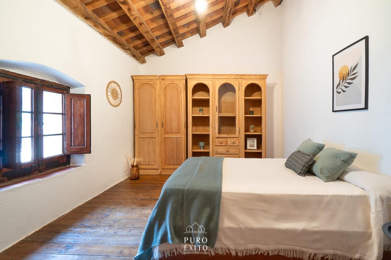 A bedroom with a double bed, wide pine wardrobe and dresser, wooden floorboards and a sloping beamed ceiling.