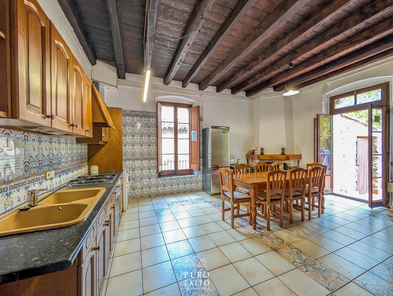 A long kitchen with patterned blue-and-white wall tiles, a gas hob, a fridge and a wooden table at the far end beneath dark beams.
